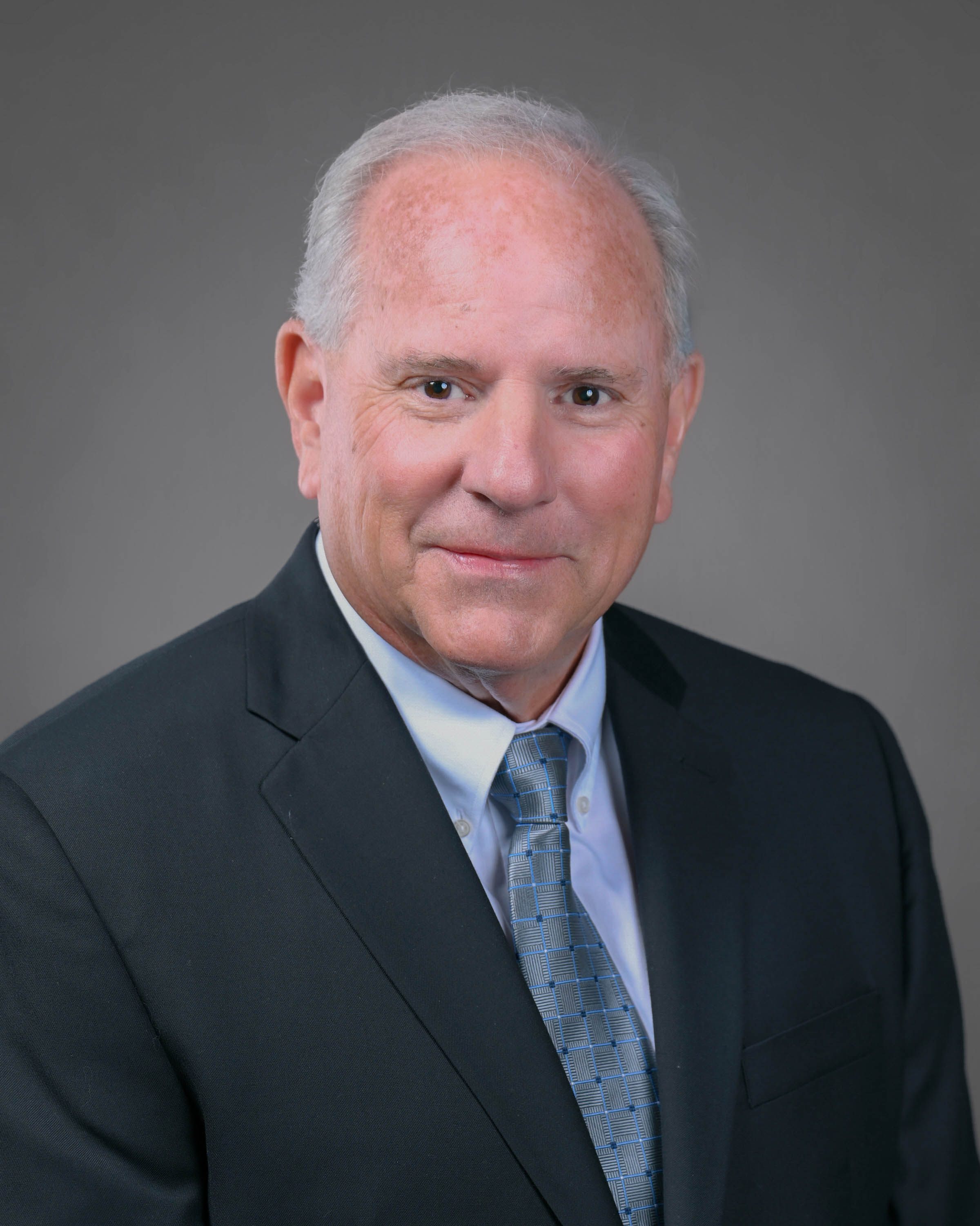 Kind man smiling with a dark grey suit and blue tie on.